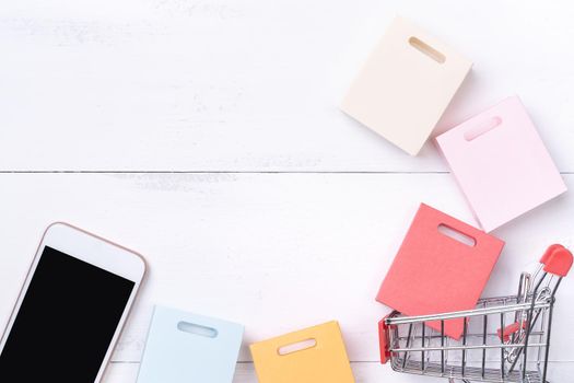 Abstract online shopping, mobile payment concept design element, colorful cart and paper bags on white wooden table background, top view, flat lay