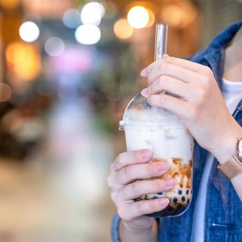 Young girl in denim jacket is drinking brown sugar flavored tapioca pearl bubble milk tea with glass straw in night market of Taiwan, close up, bokeh