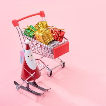 Christmas shopping concept, mini red shop cart trolley with Santa Claus toy and gift box isolated on pale pink background, blank copy space, close up