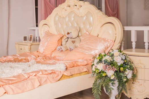 Teddy bear sitting on white bed in cozy and light bedroom.