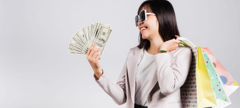 Woman with glasses confident shopper smile holding online shopping bags multicolor and dollar money banknotes on hand, excited happy Asian young female person studio shot isolated on white background