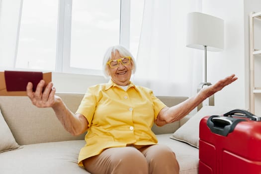 Happy senior woman with passport and travel ticket packed a red suitcase, vacation and health care. Smiling old woman joyfully sitting on the sofa before the trip raised her hands up in joy. High quality photo