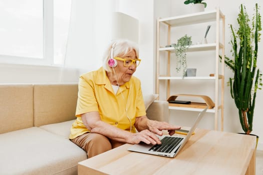 Happy elderly woman with a laptop typing with headphones sitting at home on the couch in a yellow shirt, bright modern interior, lifestyle online communication. High quality photo