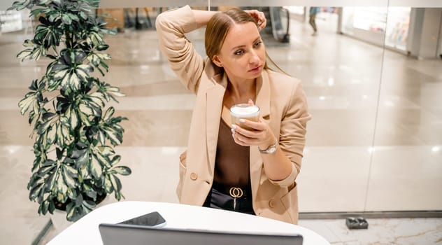 A business woman sits in a cafe, works at a computer, drinks coffee. She is wearing a beige jacket, brown top and black trousers