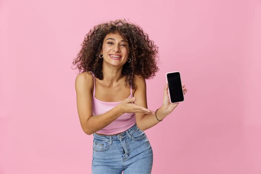 Woman blogger holding phone video call takes selfies, with curly hair in pink smile t-shirt and jeans poses on pink background, copy space, technology and social media, online. High quality photo