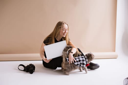 A business woman freelancer in a black is working, typing on a laptop apple and sitting on a white floor. blonde assistant in video production with computer MacBook and camera with cat