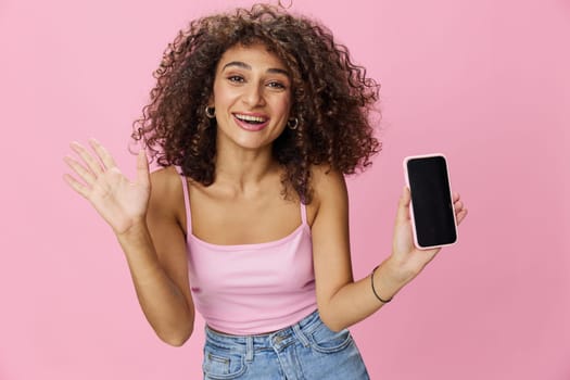 Woman blogger holding phone video call takes selfies, with curly hair in pink smile t-shirt and jeans poses on pink background, copy space, technology and social media, online. High quality photo