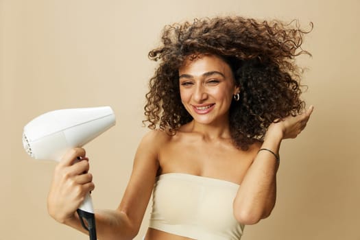 Woman dries curly hair with blow dryer, home beats styling products, smile on beige background. High quality photo