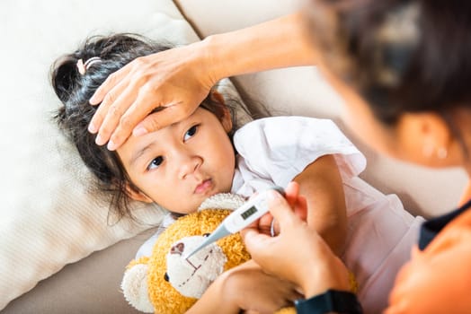 Sick kid. Mother checking temperature of her sick daughter with thermometer in mouth, child laying in bed taking measuring her temperature for fever and illness