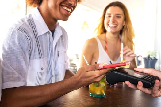 Close-up of young African American man paying bill at beach bar with phone. Black male paying round of cocktail drinks with mobile phone on summer vacation. Technology and leisure concept.