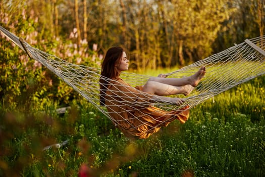 a joyful woman is sitting in a mesh hammock in nature relaxing and enjoying the rays of the setting sun on a warm summer day. Horizontal photo on the theme of outdoor recreation. High quality photo