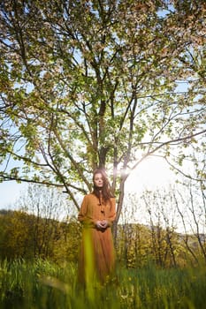 a sweet, modest woman with long hair stands in the countryside near a flowering tree and looks at the camera with her hands clasped together. High quality photo