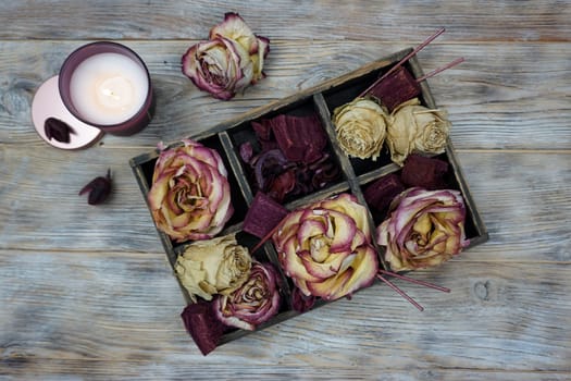 A wooden box is on the table, dried rose flowers are in the box. There is a large burning candle on the table. The concept of home comfort, hugge, self-care.