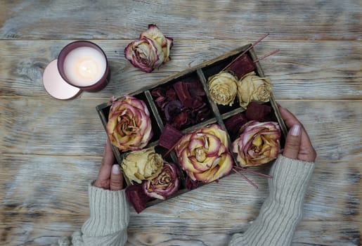 A wooden box is on the table, dried rose flowers are in the box. There is a large burning candle on the table. The concept of home comfort, hugge, self-care.