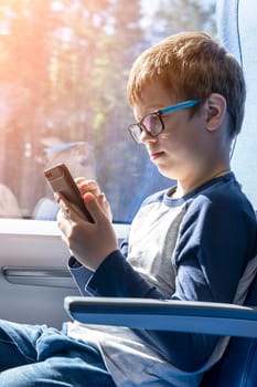 a cute Caucasian school-age boy with glasses rides a commuter train, sitting at the window. he watches cartoons on his smartphone. A fun trip. Have fun while traveling on public transport. Holidays, trip to school. vertical photo