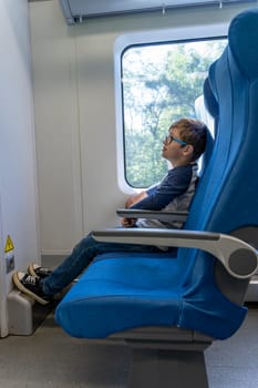 a cute Caucasian school-age boy with glasses rides a commuter train, sitting at the window. A fun trip. Have fun while traveling on public transport. Holidays, trip to school