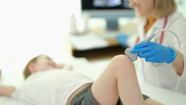 Doctor traumatologist making ultrasound examination of knee joint to child in clinic closeup. Diseases of bones and joints in children concept