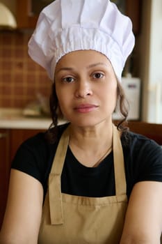 Confident portrait of a beautiful multi ethnic woman housewife, female chef baker confectioner in white hat and beige apron, confidently looking at camera. People. Profession. Career. Hobby, Lifestyle