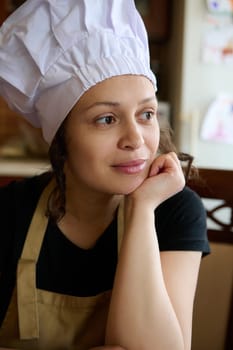 Confident authentic portrait of a beautiful multi ethnic woman, female chef confectioner, wearing white chef's hat and beige apron, dreamily looking aside. Chef cook. People. Profession. Career. Hobby