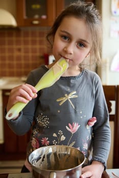 Caucasian adorable mischievous little girl 5 years old, licking utensil with chocolate sweet cream, tastes melted Belgian chocolate, standing at the kitchen table, smiling slyly looking at the camera