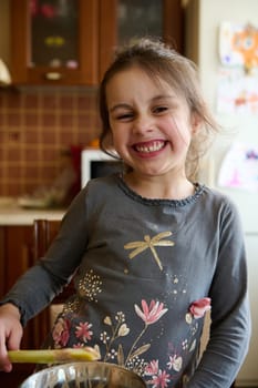 Adorable mischievous Caucasian little child girl making faces, grimacing looking at camera, tasting sweet cream whith melted Belgian chocolate, standing at the kitchen table at cozy home interior