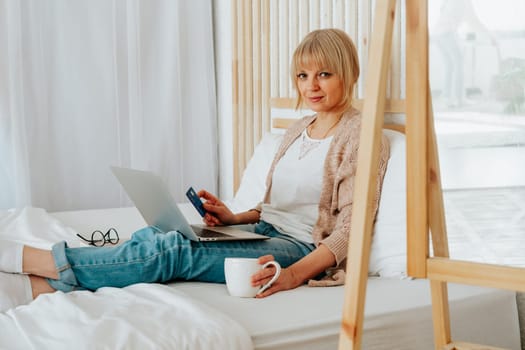 Senior mature woman sitting on bed with coffee tea mug and debit credit card while using her laptop for online shopping investments insurance.