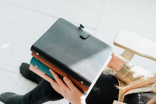 Cropped picture of young woman's hands holding stack of notebooks. Girl holds a pile of notepads books while sitting on a wooden chair. Blank space mockup books.