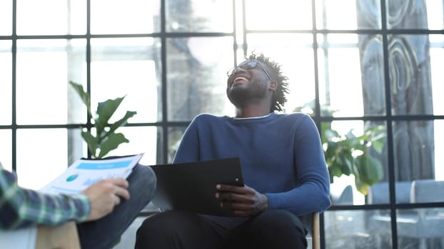 black man with a folder in his hands laughs while sitting on a chair