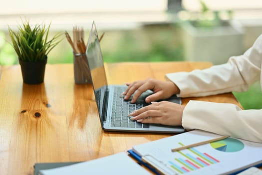 Side view of businesswoman hands typing on laptop, working with financial graphs charts online at desk.