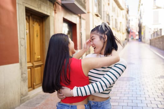 Side view of cheerful young ethnic female tourists in casual clothes standing on paved street in old town and embracing each other