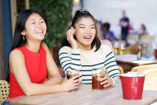 Happy young Asian female friends in casual outfits smiling and looking away while sitting at wooden table with refreshing cold beverages during meeting together