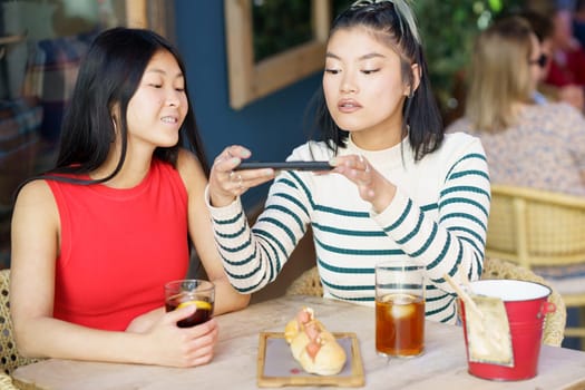 Smiling young Asian girl friends, in casual clothes sitting at table with cold drinks and taking picture of tasty food on smartphone while spending time together in cafe