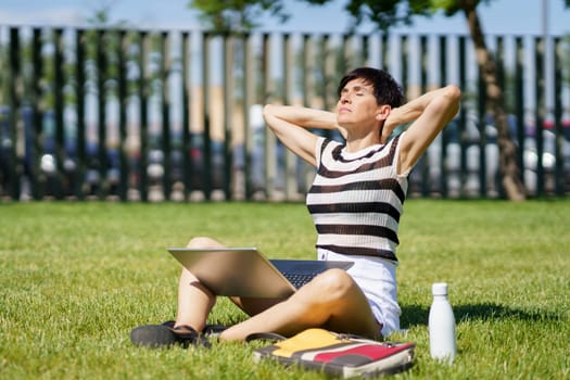 Full body of peaceful middle aged female freelancer, in summer clothes sitting on grass with laptop and closed eyes while stretching neck during remote work on project