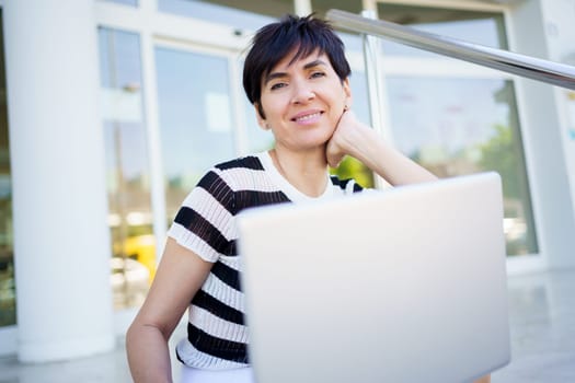 Cheerful adult female freelancer in striped t shirt, smiling and looking at camera while leaning on hand and working on remote project on laptop on street