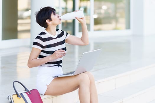 Concentrated adult female freelancer in casual summer outfit sitting on stairs and drinking water from bottle while working remotely on laptop on street
