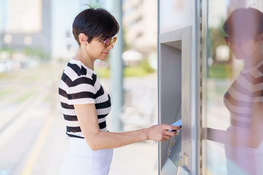 Side view of concentrated adult female in casual clothes and sunglasses paying for order with smartphone while buying ticket on terminal