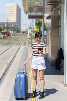 Full body of cheerful young female traveler in casual clothes and sunglasses smiling and messaging via smartphone while standing with suitcase during summer trip