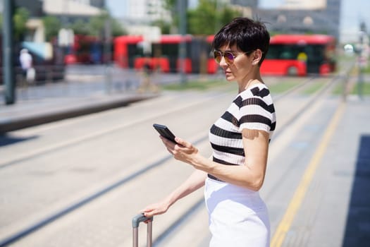 Concentrated adult female tourist in casual summer clothes and sunglasses with suitcase checking messages on mobile phone while waiting for transport