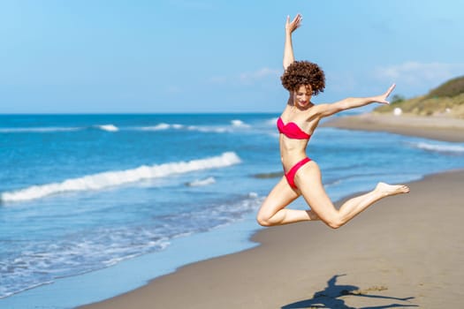 Side view of self assured young fit female, with brown curly hair in red bikini jumping with raised arms and looking down on sandy beach near wavy sea