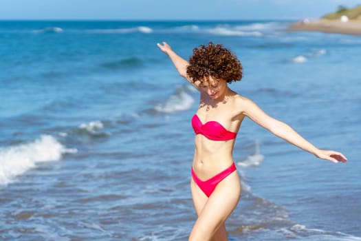 Delighted young fit female with dark curly hair, in red bikini smiling and looking down while walking in wavy sea with outstretched arms during summer holidays holidays