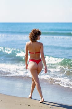 Back view of gorgeous young fit female with brown curly hair in red bikini, smiling and admiring wavy sea while strolling on sandy beach on sunny day