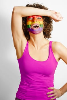 Cheerful female wearing purple tank top standing with hand on waist and painted in rainbow colors face looking at camera against white background