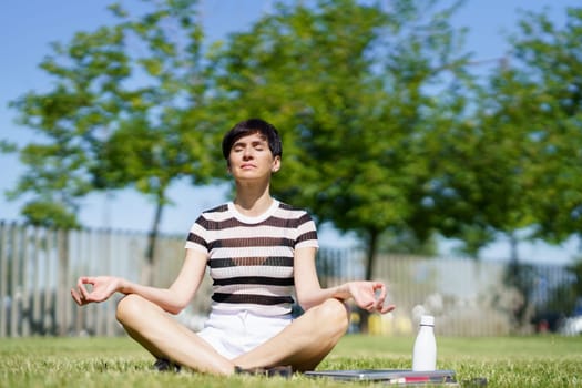 Full body of relaxed adult female in striped t shirt, sitting on grassy lawn in lotus pose with closed eyes and Gyan mudra hands while meditating during yoga session