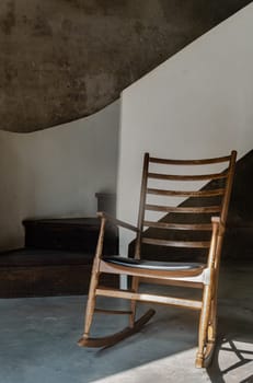 The Wooden Rocking Chair is standing alone in front of the White Spiral Staircase and Bare Cement Wall with Sunlight Shining Through. Space for text, Selective focus.