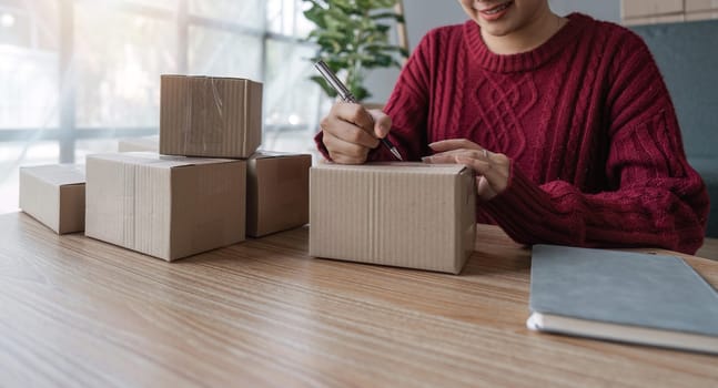 Young business woman working online e-commerce shopping at her shop. Young woman seller prepare parcel box of product for deliver to customer. Online selling, e-commerce.