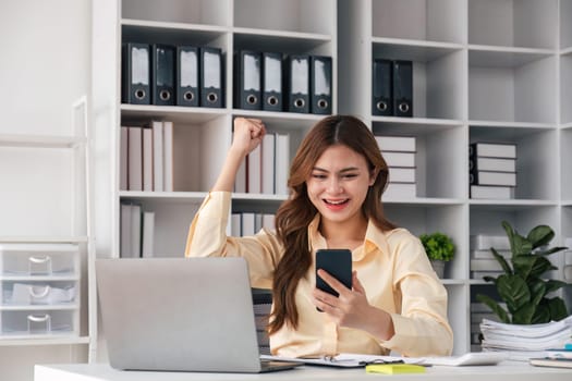 Excited businesswoman winning after achievement reading a smart phone sitting in a desk at office.