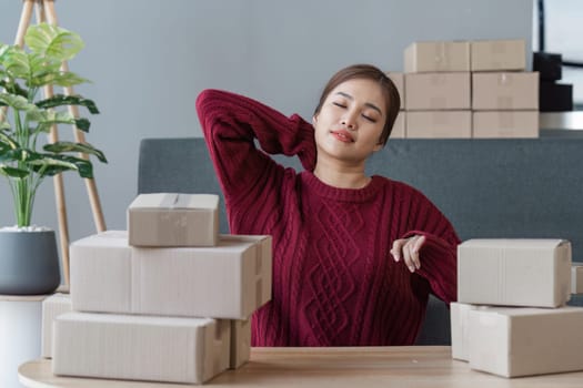 Small online business owner Asian woman stressed headache taking notes with parcel box at home.