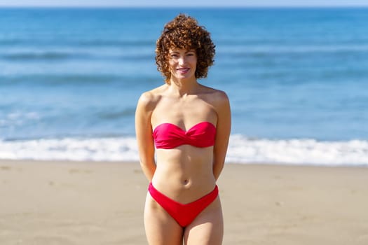 Delighted curly haired female in red swimwear standing on sandy shore against rippling sea and looking at camera in sunlight