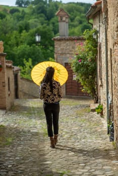 Young woman with dreadlocks braided hairstyle holding umbrella parasol. Happy young woman feeling confident in her style. Fashionable woman standing in the street of old village against stone wall.