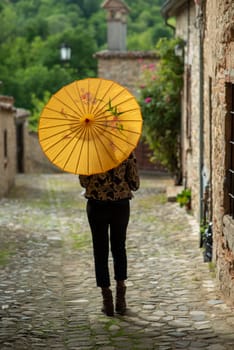 Young woman with dreadlocks braided hairstyle holding umbrella parasol. Happy young woman feeling confident in her style. Fashionable woman standing in the street of old village against stone wall.